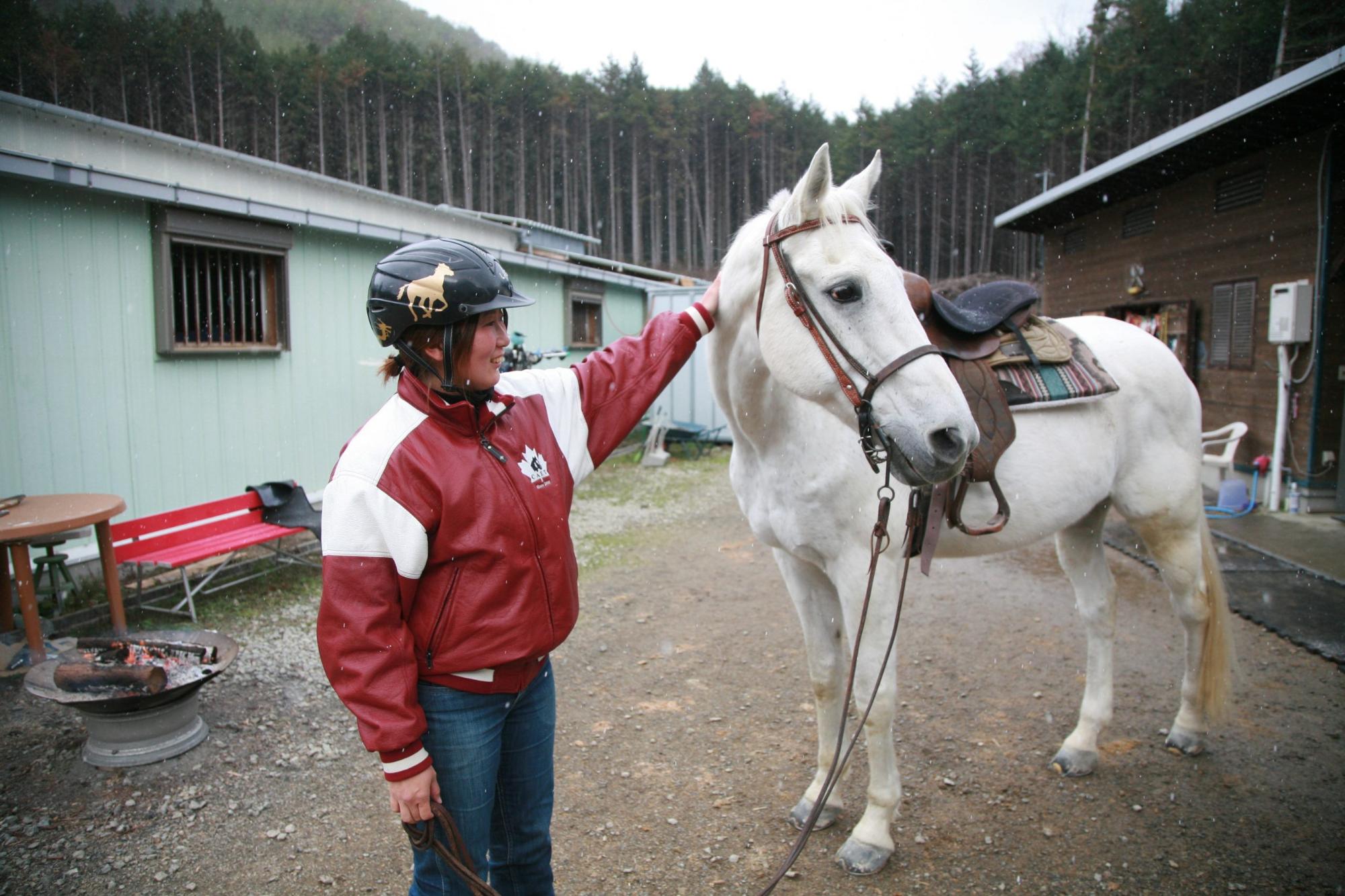 赤いジャンバーと黒いヘルメットを着用し、左に首を向けた白馬のタテガミを撫でている中山美香さんの写真