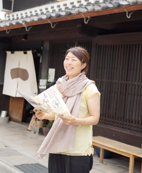 昔ながらの長屋を背景に、パンフレットを持ってほほ笑む関山 舞（せきやま まい）さんの写真