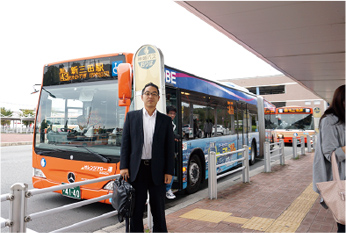 西日本初の連節バス「オレンジアロー連」の前でほほ笑むスーツ姿の男性の写真