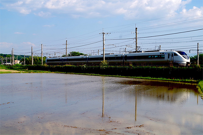 水が入っている田んぼの前を左から右へと走っていく電車の写真