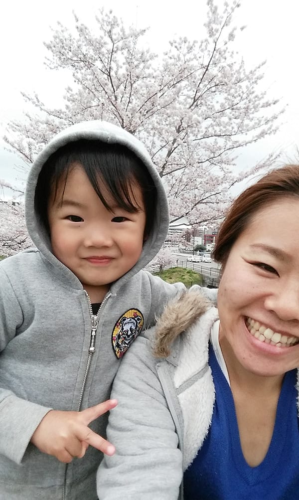 桜をバックに母親に抱かれているフードつきの洋服を着ている男の子の写真