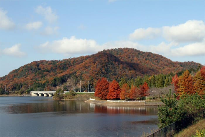 湖のバックに赤く色づく紅葉とその背後の山々の写真
