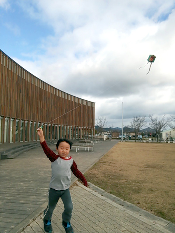 えんじ色と灰色の長袖シャツを着た児童が凧をあげている写真